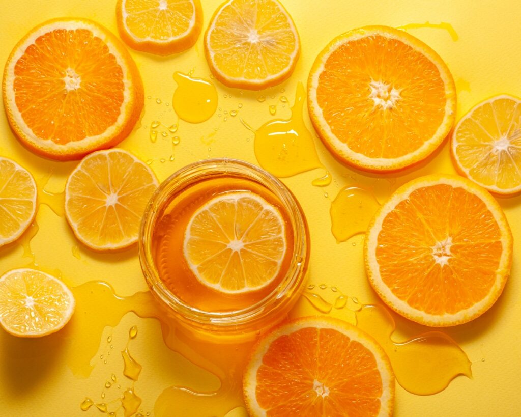 Rodelas de laranja e limão em um fundo amarelo com gotas de suco, simbolizando os benefícios da vitamina C para a pele