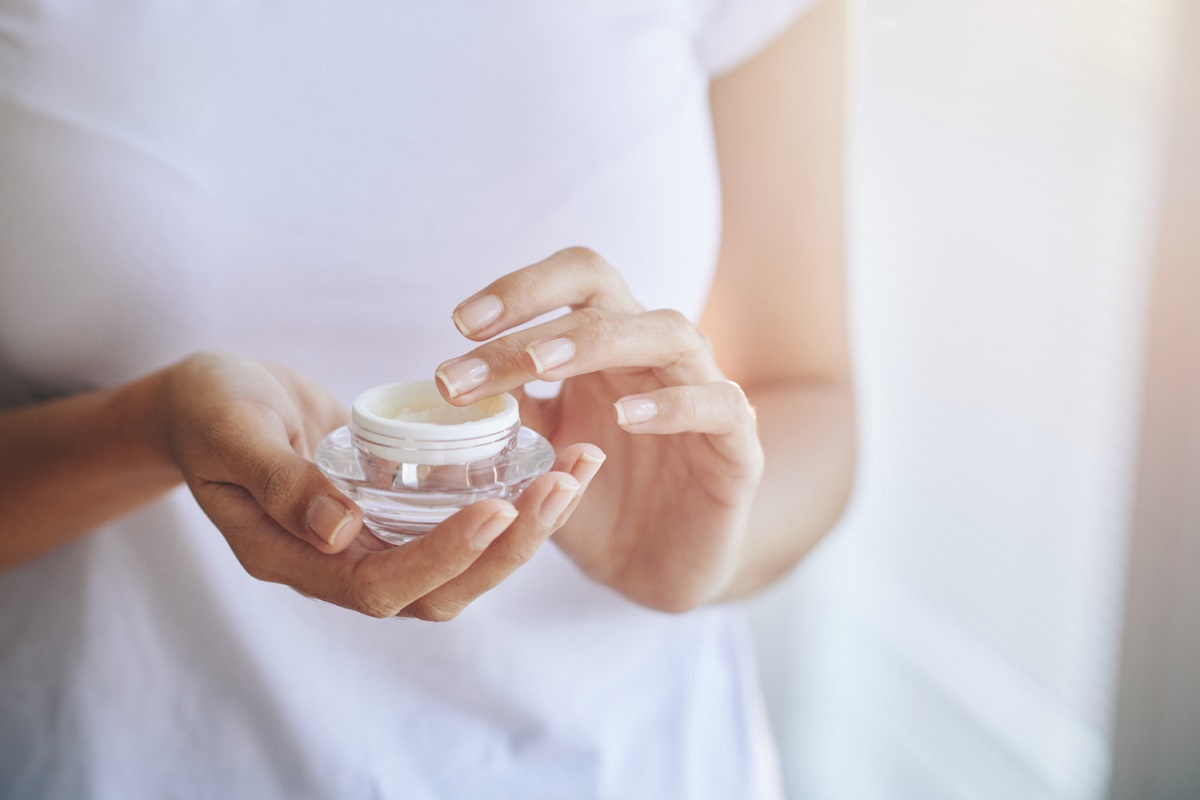 Mão de uma pessoa aplicando creme anti-idade em pote transparente, simbolizando o uso de peptídeos biomiméticos na rotina de skincare.