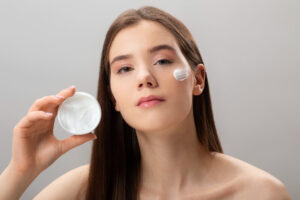 Mulher segurando um pote de creme dermocosmético e aplicando o produto no rosto para cuidado com a pele e redução de manchas